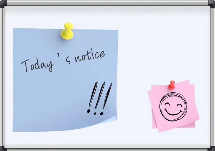 A white soft notice board in a silver aluminium frame. A blue note reading Today's notice is held by a yellow push pin, and a pink note with a drawn smiley face by a red one.