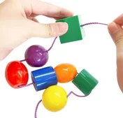 Two hands threading a large green cube bead onto a cord, with round, oval and cylindrical beads in purple, red, blue, orange, yellow and green below.