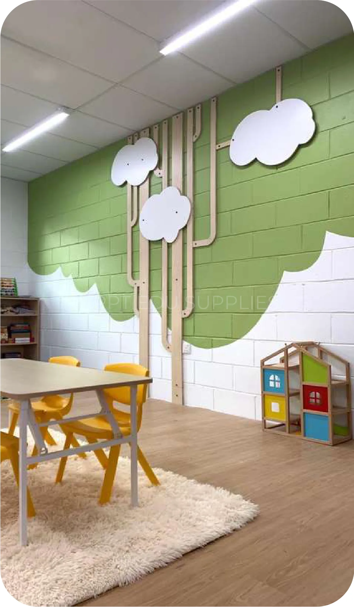 A classroom with a green wall painted with white clouds, a round table with small chairs and a pale rug.