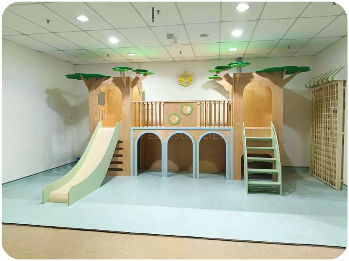 A two storey timber play frame with a rail, a stair at one end and a long slide at the other, in a pale green hospital room.
