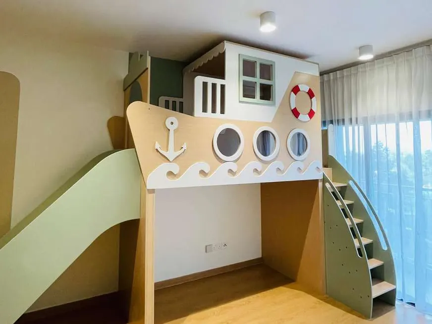 Boat-shaped plywood loft playhouse with anchor, portholes, lifebuoy and wave trim, a green slide and staircase, in an empty room.