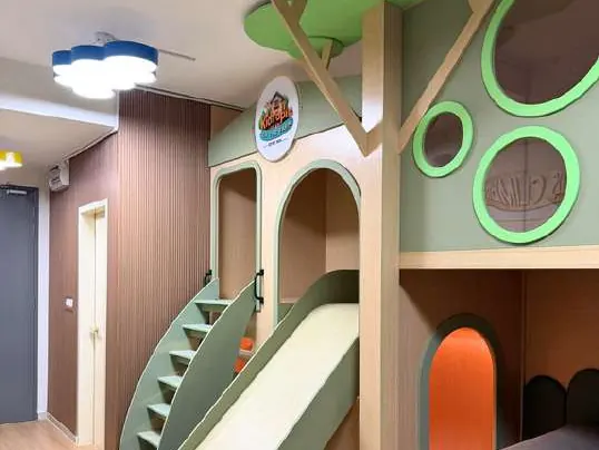 Indoor plywood treehouse with green canopies, a sage slide, staircase and arched den openings in an empty apartment playroom.