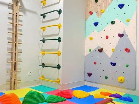 Therapy room corner with timber wall bars, a pegboard climbing wall painted as mountains, foam floor tiles and stepping stones.
