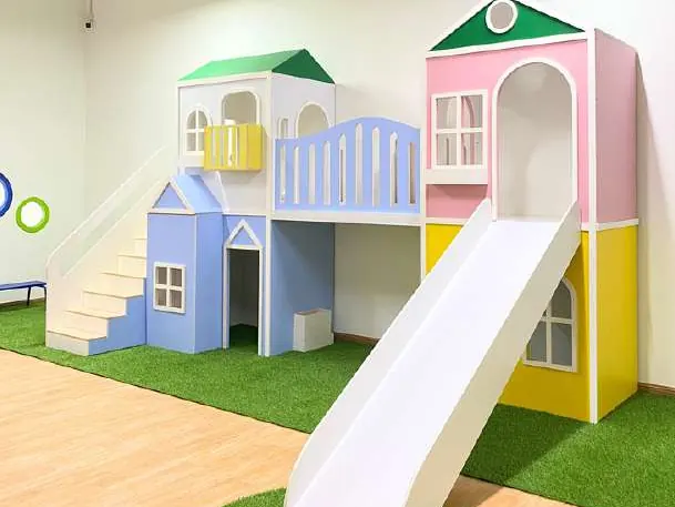 Two-storey playhouse in pastel blue, pink and yellow with green roofs, a white slide, and artificial grass on the floor.