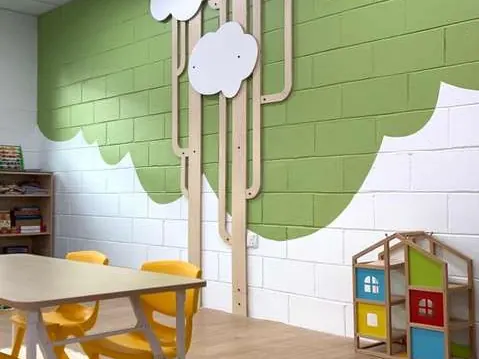 Classroom with a green block wall, plywood cloud panels on tall stems, yellow chairs at a table, and a house-shaped shelf.