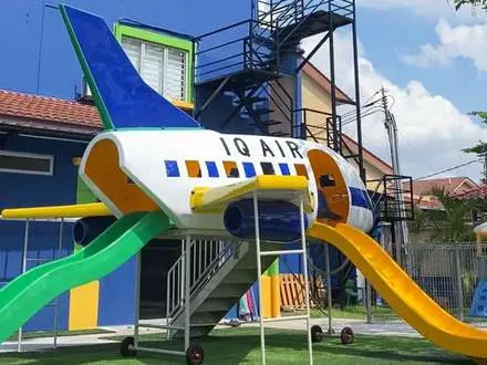 Outdoor aeroplane playground lettered IQ AIR, with green and yellow slides, on artificial turf beside a blue two-storey kindergarten building.